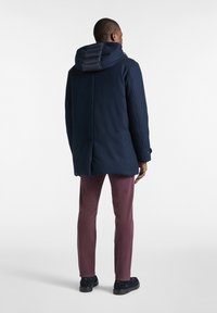 Mornar blue kaput s punjenim kapuljačom, elegantnog dizajna i dugmadima kao ukras. Nošen preko burgundskih hlača i crnih cipela.