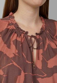 Blouse brun et terracotta à motif, avec un col froncé, présentant une ouverture en forme de clé et un lien de fermeture à l'avant.