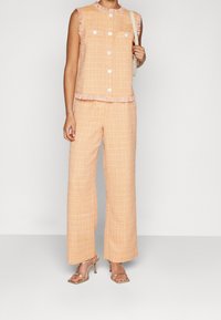 Brzoskwiniowa kamizelka z tweedu z frędzlami na rękawach i przednimi kieszeniami, połączona z szerokimi spodniami. Metaliczne sandały na obcasie dopełniają stylizację.