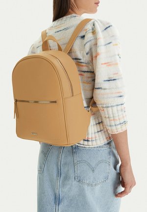 Donna con zaino in pelle color cuoio con tasca con cerniera, in piedi con maglione multicolore a maglia e jeans di denim azzurro chiaro.