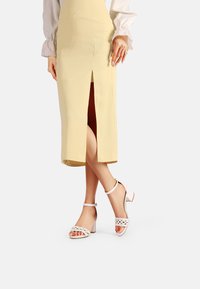 Gonna gialla al ginocchio con spacco frontale, abbinata a sandali bianchi con tacco caratterizzati da dettagli perforati e cinturini alla caviglia.