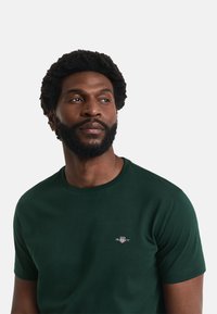 Tricou din bumbac de culoare verde închis, cu guler rotund texturat, mâneci scurte și un mic logo brodat pe piept. Textură netedă.