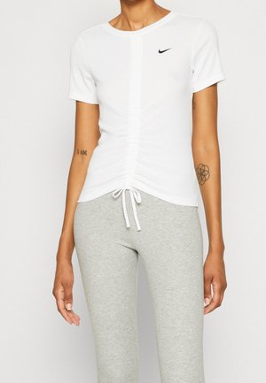 Eng anliegendes weißes Nike-Oberteil mit gerafften Ärmeln und Kordelzug, kombiniert mit hellgrauen, figurbetonten Hosen auf einer Person mit sichtbaren Arm-Tattoos.