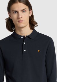 Marineblauwe polo met lange mouwen, gemaakt van ademende stof, met een knoopsluiting op de kraag en een borduursel van een oranje logo op de linkerborst.
