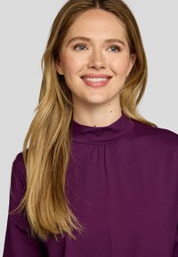 Langärmeliges lila Oberteil mit hohem Kragen, aus einem weichen Stoff, mit dezenten Falten rund um den Kragen und einer glatten Textur.