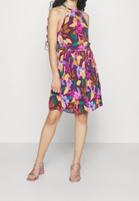 Woman wearing a sleeveless purple dress with multicolored floral print, tied waist belt, and pink strappy block-heeled sandals.