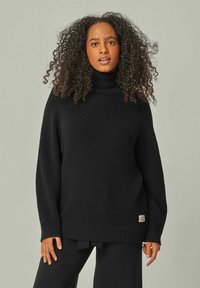 Schwarzer Strickpullover mit Rollkragen, gerippter Textur, lockerer Passform, überschnittenen Schultern und einem kleinen Logo-Patch am Saum.