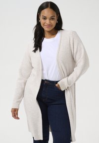 Cardigan lungo beige con una morbida texture a maglia, apertura frontale e polsini a costine, indossato sopra una t-shirt bianca e jeans blu scuro.