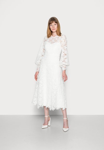 Rochie albă din dantelă cu broderie florală, mâneci lungi din tul și fustă evazată până la genunchi. Asortată cu sandale albe cu curea la gleznă.