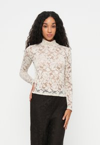 Top in pizzo a maniche lunghe color crema con colletto alto. Motivo floreale su tutta la superficie, semi-trasparente, abbinato a pantaloni neri texturizzati.