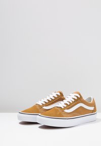 Tan suede sneakers met witte veters en zijstrepen, met een platte witte rubberen zool en een klassiek ontwerp met eenvoudige stikdetails.