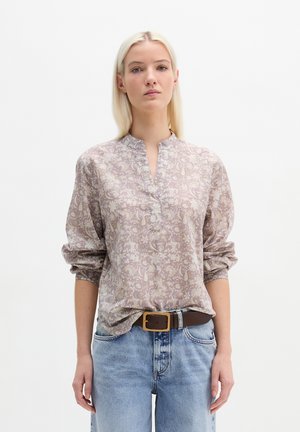 Blonde vrouw draagt een lichtpaarse blouse met bloemmotief en opgerolde mouwen, ingestopt in lichtblauwe spijkerbroek, staand tegen een witte achtergrond.