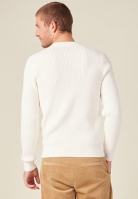 Weißer, strukturierter Pullover mit Waffelpattan, gerippten Bündchen und Saum. Modell von hinten gezeigt, kombiniert mit hellbraunen Cordhose.
