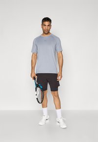 Lys grå kortermet treningsshirt med en subtil logo, parret med svarte shorts. Modellen holder en tennisracket og har på seg hvite joggesko.