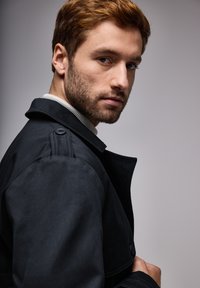 Abrigo negro de trench con un collar estructurado, detalles de botones en los hombros, usado sobre un suéter gris claro. Tejido suave, diseño ajustado.