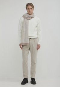 Giovane uomo in piedi che indossa un maglione bianco, pantaloni in velluto a coste beige, una sciarpa beige a righe e scarpe eleganti nere, su uno sfondo semplice.