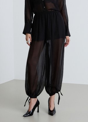 Woman wearing sheer black pants with drawstring cuffs, black high heels, and a sheer long-sleeve blouse standing on a gray floor.