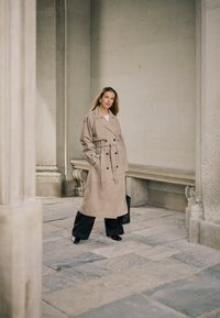 Kobieta w beżowym trenczu i czarnych spodniach stoi na kamiennym dziedzińcu z klasycznymi kolumnami i ławką, trzyma czarną torbę.