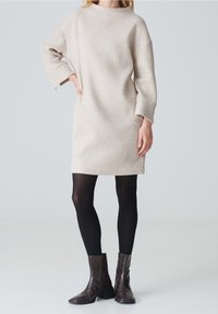 Robe beige en mélange de laine, coupe ample, encolure bateau et manches longues ; associée à des collants noirs et des bottines marron.