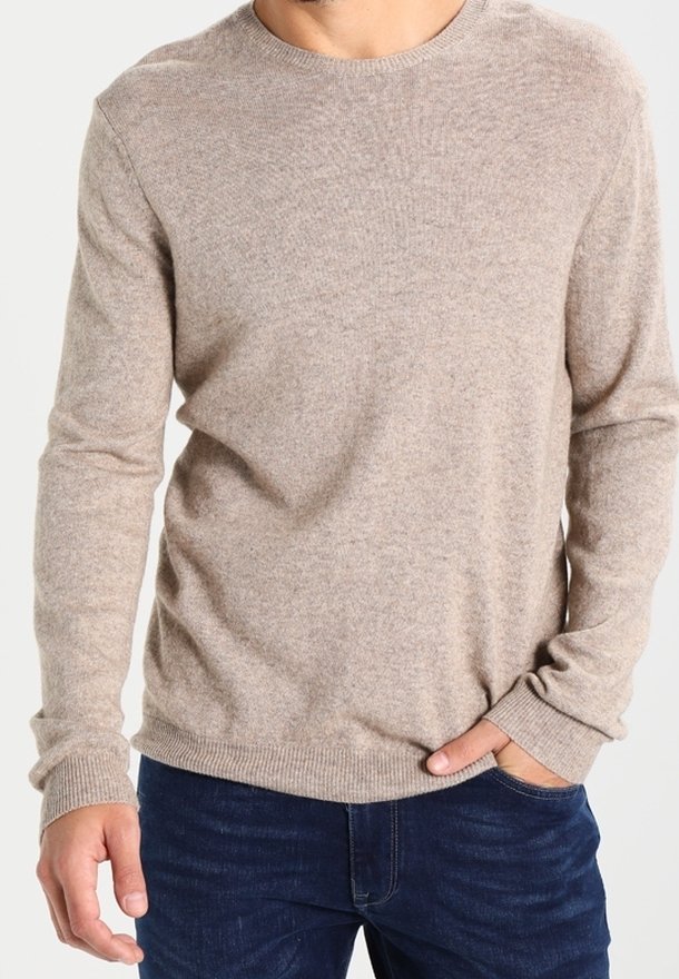 Pull à manches longues beige à col rond porté avec un jean bleu foncé, une main dans la poche, sur un fond uni.