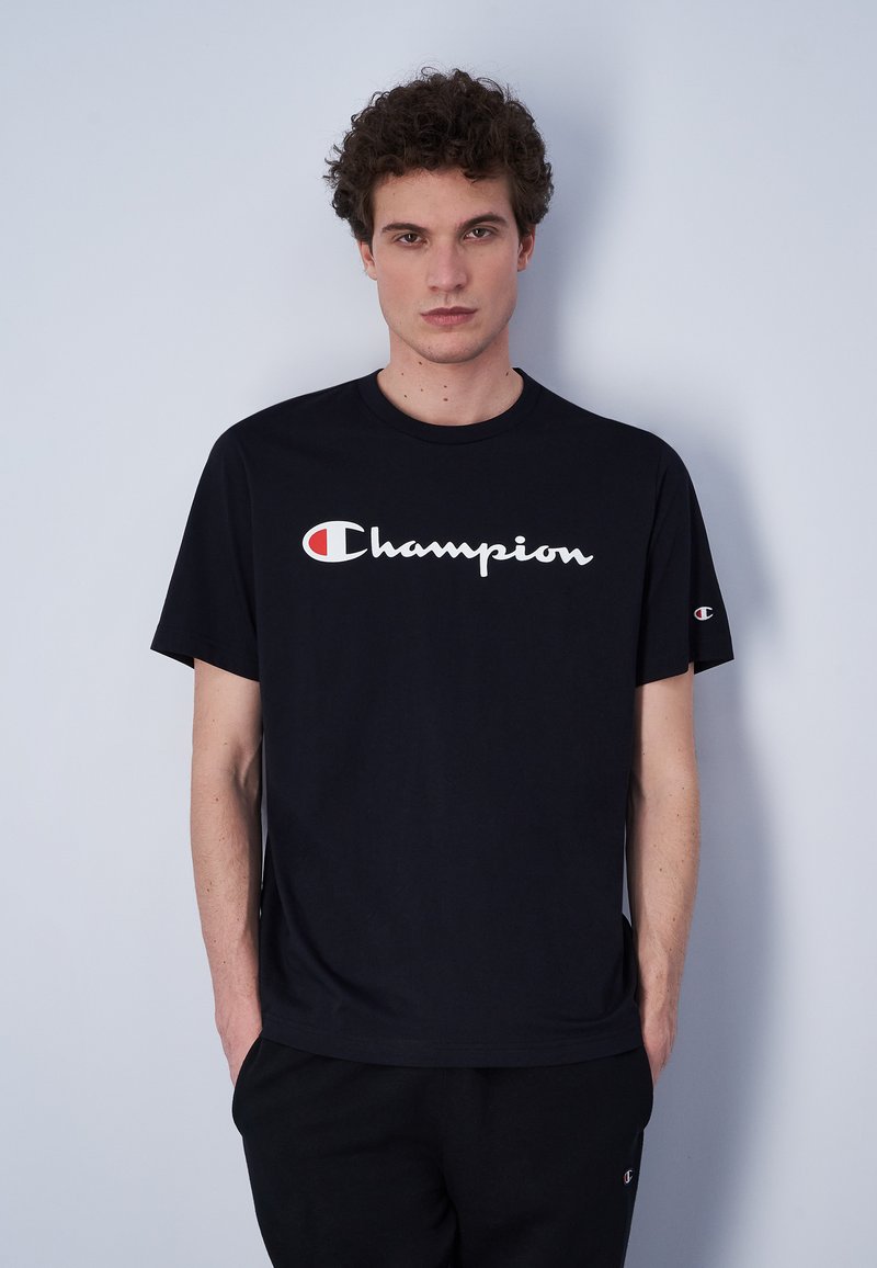 Camiseta de algodón negra con un gran logo blanco de "Champion" con acentos rojos. Mangas cortas y un cuello redondo clásico.