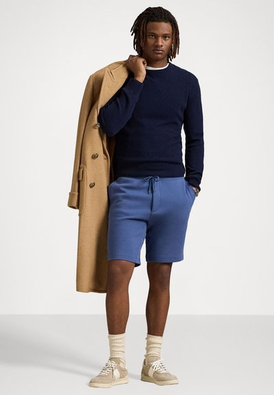 Pulover bleumarin, pantaloni scurți albaștri, palton bej drapat pe umăr. Tenisi bej, șosete crem. Textură netedă, fit casual, design minimal.