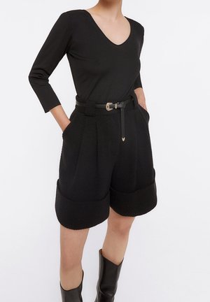 Donna che indossa una maglia nera con scollo a V, pantaloncini a vita alta con texture e ampi polsini, cintura nera e stivali neri fino al ginocchio, in piedi con le mani in tasca.