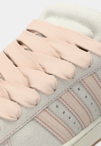 Ανοιχτό γκρι σουέντ αθλητικό παπούτσι με ροζ υφασμένες κορδόνες, τρεις ροζ ρίγες στο πλάι και διακριτικές ραφές στην επάνω επιφάνεια.