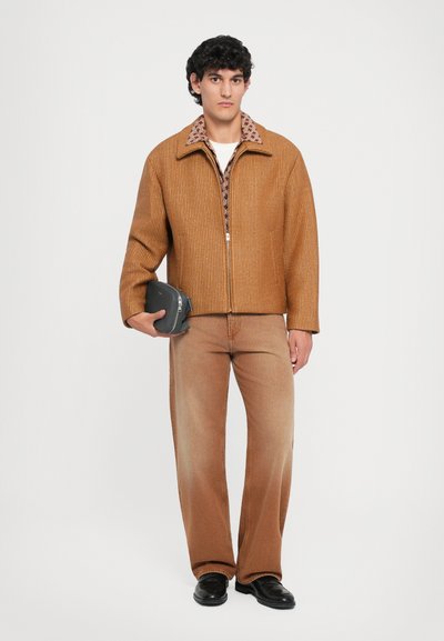 Chaqueta marrón de canalé con cremallera y cuello estampado, combinada con jeans anchos de un marrón desvaído y zapatos negros, sosteniendo un bolso pequeño oscuro.
