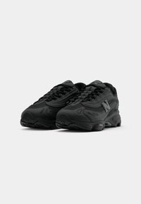 Des chaussures de sport noires avec une tige synthétique texturée, un système de laçage et une semelle rembourrée. Elles présentent un logo réfléchissant sur le côté.