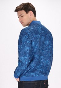 Giacca bomber blu con motivo a foglie, colletto e polsini a coste, con una texture liscia e un tessuto leggero.