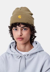 Gorro de punto verde oliva con un logo amarillo en la parte frontal, llevado con una sudadera ligera gris claro que presenta un detalle de cordón.
