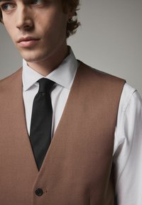 Gilet texturé marron avec une cravate noire sur une chemise blanche, comportant un seul bouton et des détails de col prononcés. Vue en gros plan.