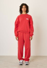 Punane puuvillane dressipluus langetatud õlgade ja ümarate kaelustega, sobitatud sama punase puuvillase dressipüksiga. Hallid spordijalanõud.