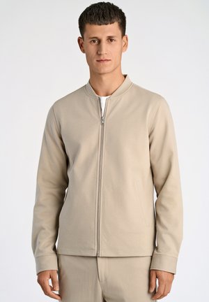Beige ritjack met een ronde halslijn, lange mouwen en zijzakken, gemaakt van een zacht materiaal. Minimalistisch ontwerp.