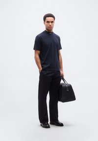 Μπλε σκούρο κοντομάνικο T-shirt με υψηλό γιακά και λεπτομέρεια λογότυπου· μαύρο παντελόνι ραφής· κρατώντας μαύρη δερμάτινη τσάντα· μαύρα παπούτσια.