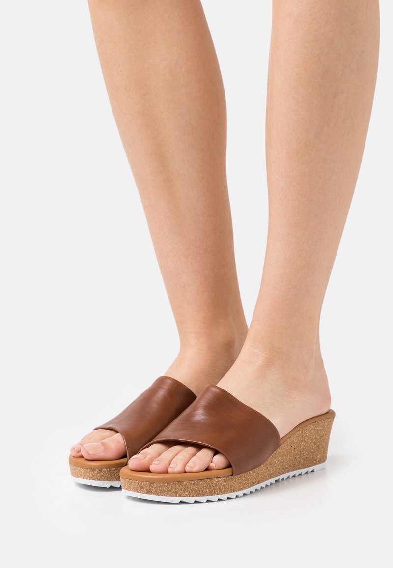 Sandales compensées en cuir marron avec une semelle en liège et des accents en caoutchouc blanc. Design ouvert à l'avant, texture lisse et légère courbe au niveau du talon.