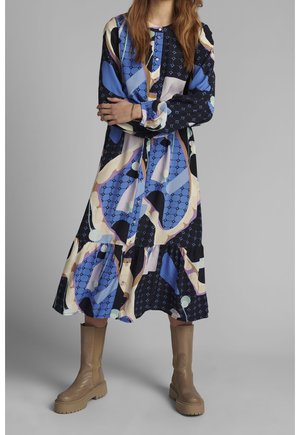 Kobieta w kolorowej sukience midi z abstrakcyjnym wzorem, z długimi rękawami, oraz beżowych, masywnych botkach sięgających do połowy łydki, stoi ze skrzyżowanymi ramionami.