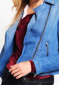Chaqueta de mezclilla en azul claro con cierre frontal, bolsillo con cremallera y cuello estructurado. Llevada sobre una blusa burdeos de manga larga.