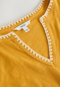 Vêtement jaune moutarde avec bordure en crochet triangulaire blanc le long de l'encolure en V et du col intérieur, avec une étiquette de marque "NEXT" et une étiquette "Made in India".