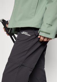 Hand houdt een zwarte wandelstokriem vast, draagt een lichtgroene jas met TEXAPORE-tekst en zwarte Jack Wolfskin broek met zichtbaar logo.