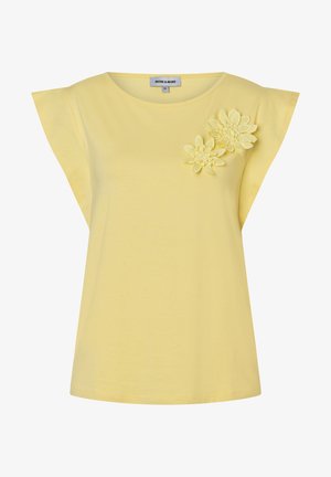 Gelbe Bluse mit kurzen Ärmeln und weitwinkligen Ärmeln sowie zwei strukturierten Blumenapplikationen auf der oberen linken Brustseite.