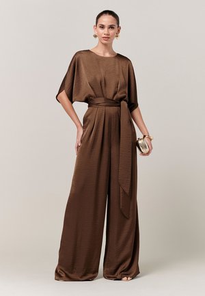 Femme portant une combinaison pantalon marron à jambes larges avec une ceinture nouée à la taille, tenant une pochette dorée et portant des boucles d'oreilles créoles dorées ainsi qu'un bracelet doré.