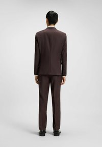 Brown tailored suit made of textured fabric; features slim fit jacket and matching trousers, with black shoes visible. No visible patterns.