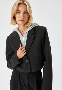 Blazer negro corto con un solo botón, usado sobre una sudadera con cremallera gris claro. Presenta mangas anchas y una textura de tela suave.