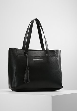 Černá kožená taška tote s dvojitými uchy a ozdobou třásní, umístěná na bílém podstavci na jednobarevném světle šedém pozadí.