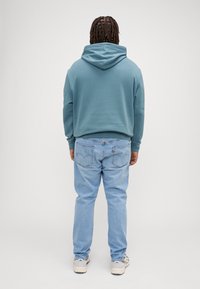 Sweatshirt capuz verde-azulado com um corte descontraído, combinado com jeans azul-claro. Detalhes visíveis nos bolsos traseiros com a marca Levi's.