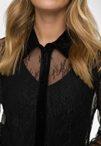 Mustame bluus, millel on läbipaistvad pitsi- ja võrkpaneelid. Kraega disain ees olevate nööpidega, tekstuurne kangas ja delikaatsed lillemustrid.