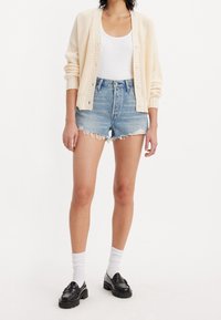 Cárdigan de punto beige claro, camiseta sin mangas blanca, pantalones cortos de denim azul deshilachados, mocasines negros y calcetas blancas.