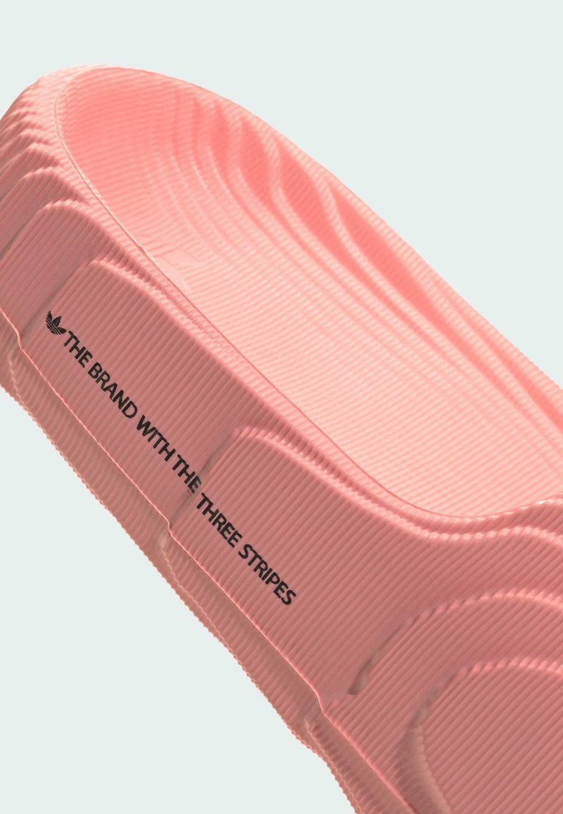 Chaussure slip-on en caoutchouc rose avec texture côtelée, arborant le texte de la marque en noir. Design incurvé et bords lisses mettant en avant la fonctionnalité.